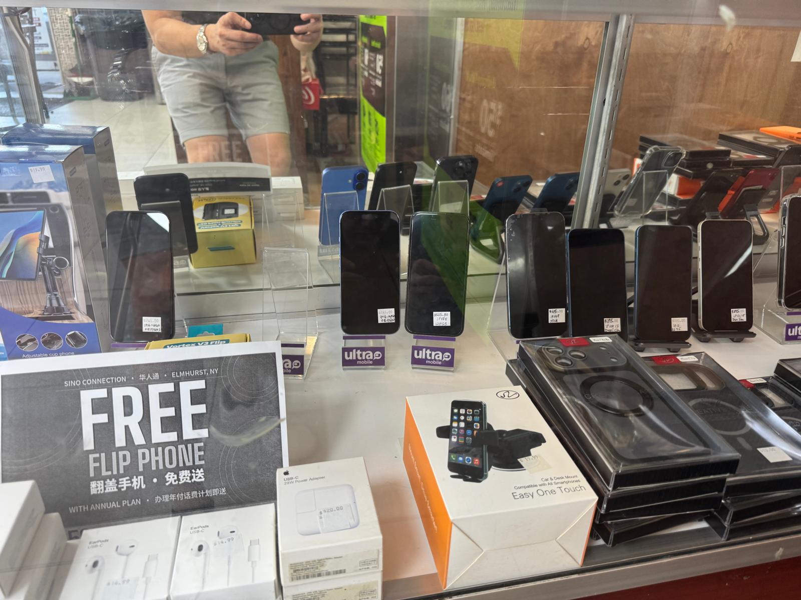 展示櫃裡的 iPhone,立在架上,每台都貼著手寫價格,還有耳機、充電器和車用支架,旁邊是「辦理年付話費計劃送摺疊手機」的牌子